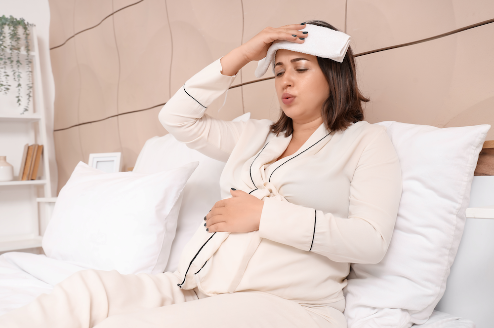 A pregnant person leans back in bed with a towel over their forehead.