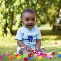 A happy toddler sits playing with blocks in a shady park.