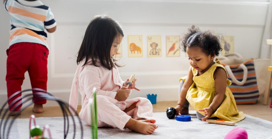 Toddlers play with toys together in a sunny play room.