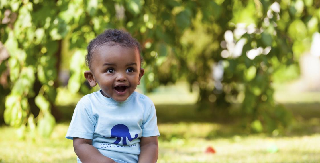 A happy toddler sits playing with blocks in a shady park.